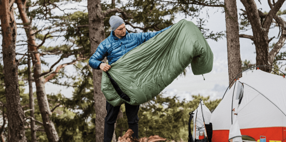 Man håller upp stor ljusgrön sovsäck vid tält i skog