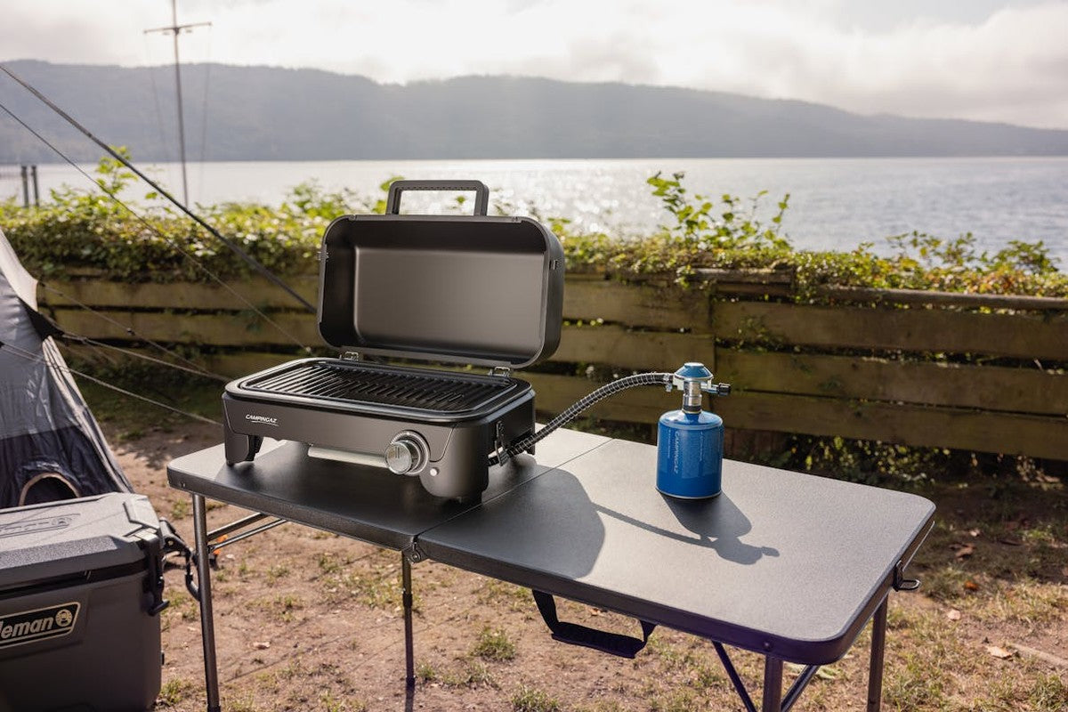 Mörkgrått campingbord med splittrad yta bär CAMPGRILL vid sjö